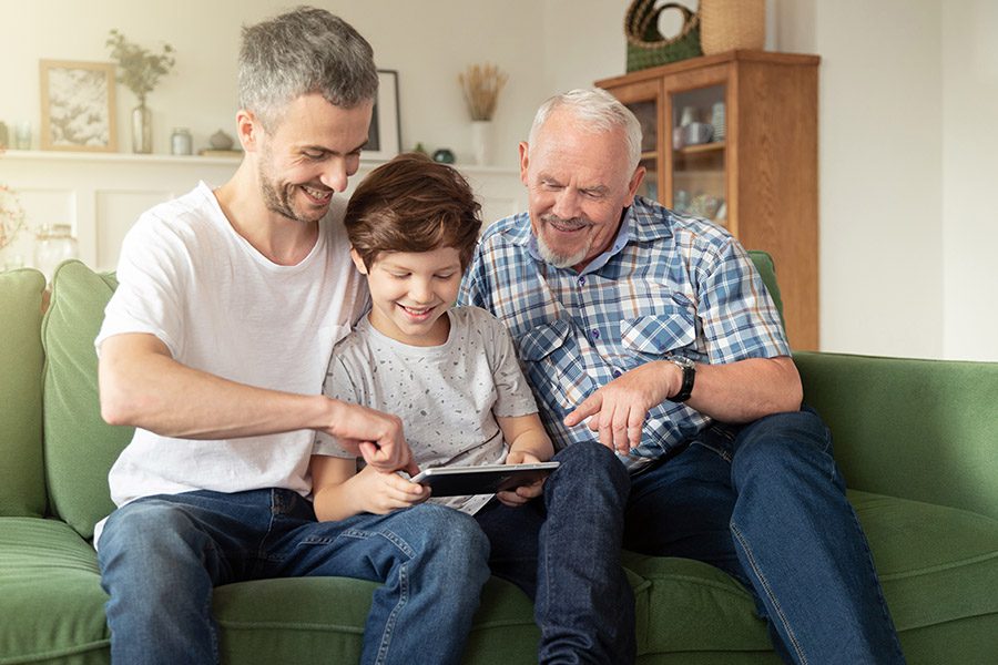 Client Center - A Grandfather, Father and Son are Sitting Together on a Sofa Smiling and Using a Tablet at Home